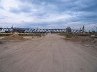 dirt road with train tracks under an overpass on the opposite track, and sand area below with vehicles and machinery