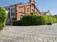 a large bricked walkway that is surrounded by bushbery and bushes, next to a brick building with red brick accents