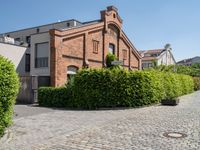 a large bricked walkway that is surrounded by bushbery and bushes, next to a brick building with red brick accents