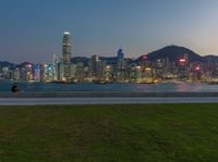 a woman is flying a kite in a city park at dusk, next to a sea and tall buildings