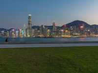 a woman is flying a kite in a city park at dusk, next to a sea and tall buildings