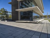 an empty bench in front of a glassed building outside a city center area with benches and trees