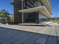 an empty bench in front of a glassed building outside a city center area with benches and trees