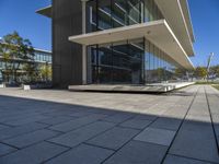 an empty bench in front of a glassed building outside a city center area with benches and trees