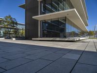 an empty bench in front of a glassed building outside a city center area with benches and trees