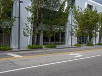 an empty street with parked cars near some tall buildings and trees in front of it