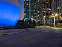 a street has some palm trees and blue lights at night in a city area with tall buildings, and an enormous balloon