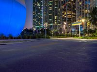 a street has some palm trees and blue lights at night in a city area with tall buildings, and an enormous balloon