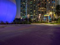 a street has some palm trees and blue lights at night in a city area with tall buildings, and an enormous balloon