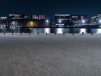 a group of benches sitting on the side of a lake next to some buildings at night