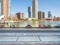 several benches and tables next to a river and a city with a bridge in front of them