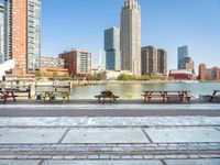 several benches and tables next to a river and a city with a bridge in front of them