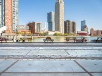 several benches and tables next to a river and a city with a bridge in front of them