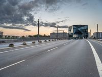 empty road with tall poles and modern glass building in background under stormy sky at dusk