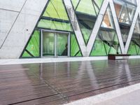 a green building with a bench in front of it on wooden floors and concrete, under glass windows