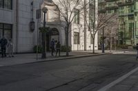 three people walking down the sidewalk on either side of a white building, some black trees and many tall buildings