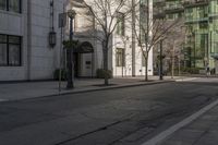 three people walking down the sidewalk on either side of a white building, some black trees and many tall buildings