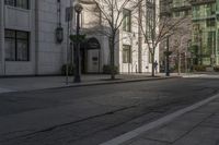 three people walking down the sidewalk on either side of a white building, some black trees and many tall buildings