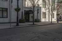 three people walking down the sidewalk on either side of a white building, some black trees and many tall buildings