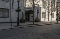 three people walking down the sidewalk on either side of a white building, some black trees and many tall buildings