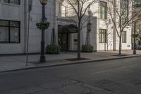 three people walking down the sidewalk on either side of a white building, some black trees and many tall buildings