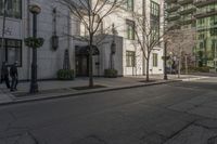 three people walking down the sidewalk on either side of a white building, some black trees and many tall buildings