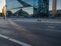 a person sits in the road alone on their motorcycle near a building that has a glass facade