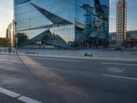 a person sits in the road alone on their motorcycle near a building that has a glass facade