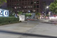 a picture of the street corner in front of a building and sign with a lot of windows, lights, bushes and trees
