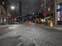 the road and street signs at night, showing a bridge in the distance with cars parked near the curb