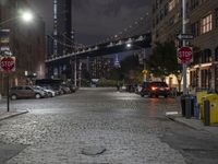 the road and street signs at night, showing a bridge in the distance with cars parked near the curb