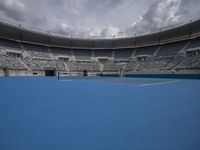 a tennis court in a stadium under a cloudy sky, with white and blue seating chairs