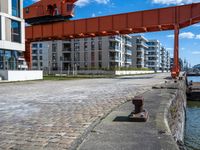 a long dock with concrete bricks beside a marina area with orange structures and buildings on the other side