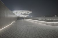 an empty walkway with lighting beside concrete walls and a building at night in the background