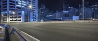 an empty parking area in front of buildings and city lights at night with a blue hued car driving past it