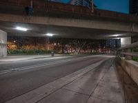 a long road under an elevated bridge with a bench on it at night and lights on