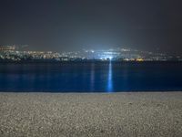 a bench sitting near the ocean during night with lights on mountains in the background on a cloudy day
