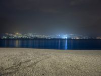 a bench sitting near the ocean during night with lights on mountains in the background on a cloudy day