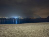 a bench sitting near the ocean during night with lights on mountains in the background on a cloudy day