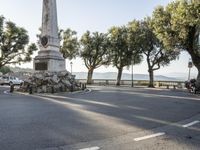 a city park with trees and large rocks and a monument on the corner, while parked cars are waiting near the curb