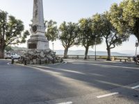 a city park with trees and large rocks and a monument on the corner, while parked cars are waiting near the curb