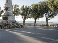 a city park with trees and large rocks and a monument on the corner, while parked cars are waiting near the curb