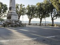 a city park with trees and large rocks and a monument on the corner, while parked cars are waiting near the curb