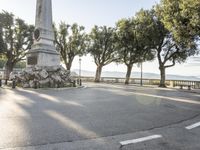 a city park with trees and large rocks and a monument on the corner, while parked cars are waiting near the curb