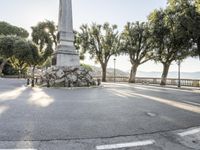 a city park with trees and large rocks and a monument on the corner, while parked cars are waiting near the curb