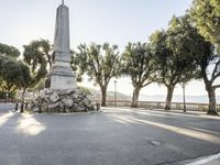 a city park with trees and large rocks and a monument on the corner, while parked cars are waiting near the curb