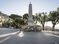 a city park with trees and large rocks and a monument on the corner, while parked cars are waiting near the curb