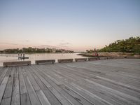 a boardwalk with benches and water in the foreground at dusk of a city by a river