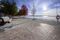 a view of trees and the sun behind a bench on a brick walkway next to the water