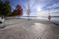 a view of trees and the sun behind a bench on a brick walkway next to the water